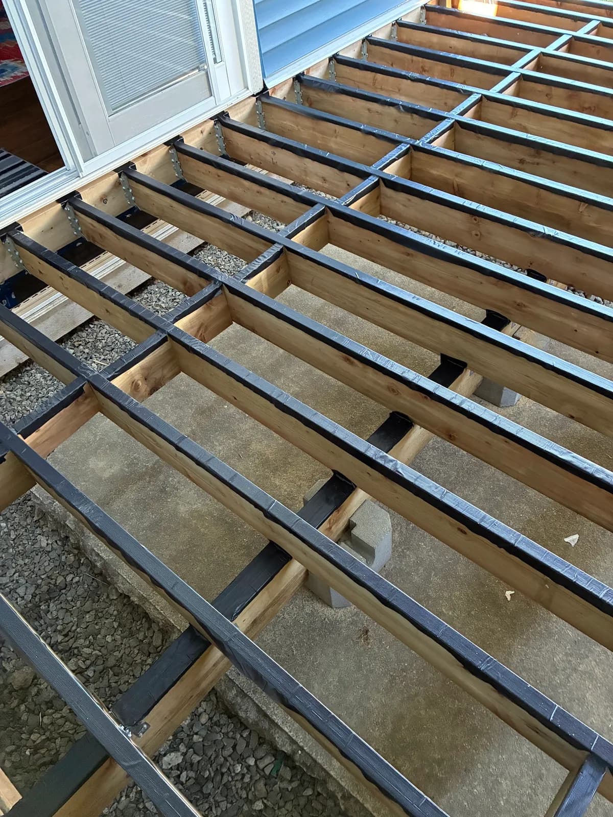 Wood deck joists and blocking with black tape along the tops beside a blue house